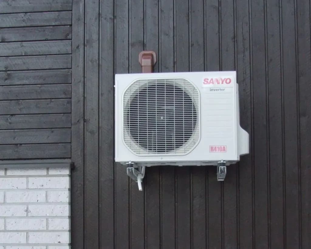 A white wall-mounted outdoor heat pump unit with a round fan grille, fixed to dark wood siding.