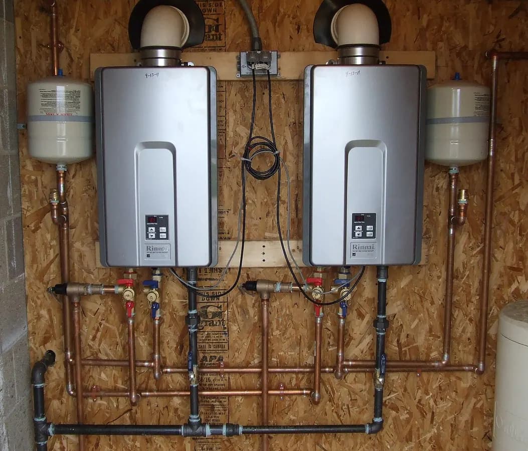 Two wall-mounted tankless water heaters with water, gas and vent piping on a wood-panel wall.